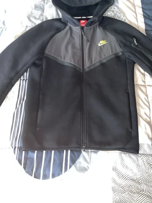 Svart Nike hoodie med dragkedja - Svart hoodie från Nike med hel dragkedja och huva. Framsidan har en grå panel upptill och Nike-logga i gult på bröstet. Ärmarna är svarta och det finns en liten ficka med dragkedja på ena ärmen. Perfekt för en sportig och avslappnad stil.