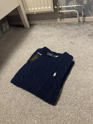 Polo Ralph Lauren stickat tröja - Mörkblå kabelstickad tröja från Polo Ralph Lauren med klassisk rund halsringning och vit broderad logga på bröstet. Tröjan har långa ärmar och ribbade muddar vid ärmslut och nederkant. Perfekt för dig som gillar stilren och tidlös design.