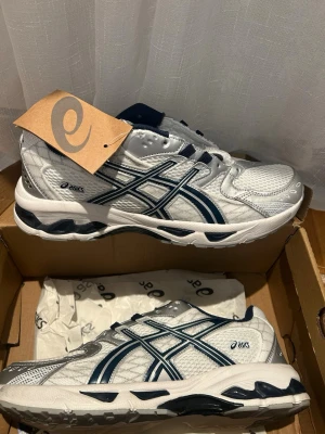 Vita och marinblå Asics sneakers - Snygga sneakers från Asics i vitt och marinblått med klassiska meshpaneler och detaljer i silver. Skorna har en låg profil, vadderad plös och häl, samt den ikoniska Asics-loggan på sidan. Perfekta för dig som gillar sportig och clean stil. Använt 2-3 gånger. 