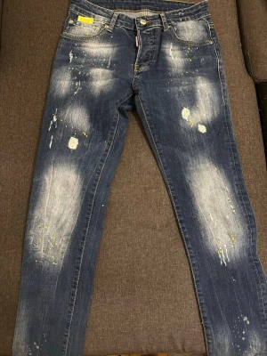Blå distressed jeans från Dsquared2 - Säljer ett par blå jeans från Dsquared2 med coolt distressed utseende, slitningar och färgstänk i vitt och gult. Klassisk femficksmodell med raka ben och låg midja. Perfekta för dig som gillar en edgy och trendig stil.
