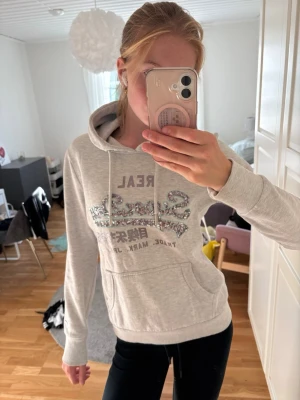 Ljusgrå hoodie med tryck - Säljer en ljusgrå hoodie med huva och dragsnören. Framtill finns ett stort tryck med text och glitterdetaljer. Hoodien har en klassisk känguruficka och ribbade muddar vid ärmslut och nederkant. Perfekt för en avslappnad och snygg stil.