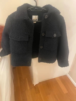 Mörk blå teddyjacka med stora fickor - Mysig mörk blå  teddyjacka med stora knappar och två rejäla bröstfickor. Jackan har en rak passform, klassisk krage och fluffigt material som ger en trendig look. Perfekt för kyliga dagar och enkel att matcha med olika outfits.