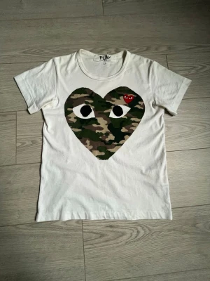 Vit t-shirt Comme des Garçons Play - ✅Vit t-shirt från Comme des Garçons Play med stort hjärta i camouflage-mönster och ögon på bröstet.✅💯 Liten röd hjärt-detalj med ögon upptill. Klassisk rund hals och korta ärmar.💯