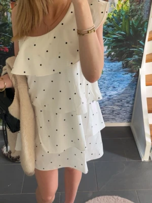 Vit volangklänning med prickar - Söt vit kortklänning med svarta prickar och volangdetaljer. Klänningen är ärmlös och har en luftig passform, perfekt för varma dagar. Styla med smycken och en liten väska för en trendig look.