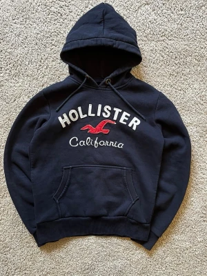 Mörkblå Hollister hoodie med huva - Snygg mörkblå hoodie från Hollister med stor broderad logga och texten 'California' på bröstet. Klassisk känguruficka framtill, ribbade muddar och justerbar huva. Perfekt för en avslappnad och trendig look.