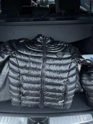 Svart pufferjacka med glansig yta - Snygg svart pufferjacka med glansig finish och quiltad design. Jackan har hög krage, två dragkedjefickor och en tydlig logga på bröstet. Perfekt för kalla vinterdagar och ger en modern look. Dragkedjan framtill har en unik detalj med logotyp.