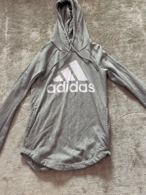 Grå hoodie från Adidas - Snygg grå hoodie från Adidas med stor vit logga på bröstet. Hoodien har dragsko i huvan, långa ärmar och en avslappnad passform. Tillverkad i mjukt material som känns skönt mot huden. Perfekt för en sportig och chill stil.
