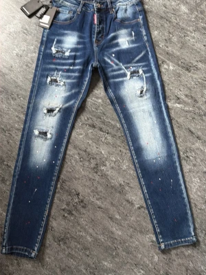 Blå slitna skinny jeans med färgstänk - Säljer ett par blå skinny jeans med slitningar och coola färgstänk i vitt och rött. Jeansen har en tvättad look med ljusare partier och trasiga detaljer på framsidan. Klassisk femficksmodell och knappgylf. Perfekta för en trendig streetstyle.