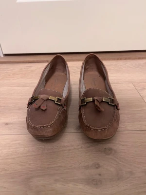 Bruna mockaloafers med metallspänne, storlek 40, Tamaris - Säljer ett par bruna loafers i mocka med dekorativt metallspänne och tofs framtill. Skorna har klassisk söm runt tån och randigt innerfoder. Perfekta för dig som gillar en stilren och avslappnad look.