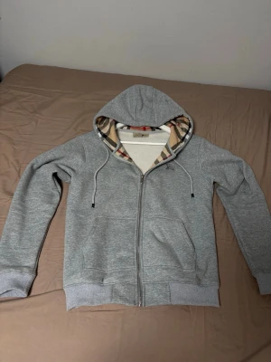 Grå zip hoodie från Burberry - Snygg grå zip hoodie från Burberry med klassiskt rutigt foder i huvan och dragsko. Mjuk bomullsblandning, två fickor fram och ribbade muddar. Perfekt för en chill och stilren look.