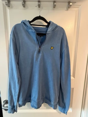 Blå hoodie från Lyle & Scott - Snygg blå hoodie från Lyle & Scott med halv dragkedja och klassisk gul logga på bröstet. Hoodien har huva och ribbade muddar vid ärmslut och nederkant. Perfekt för en avslappnad och stilren look.