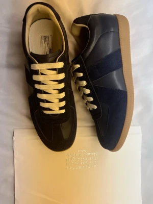 Maison Margiela - Snygga mörkblå sneakers med ovandel i mocka och läder, vita snören och klassisk gummisula i beige. Skorna har en retroinspirerad design med paneler i olika material och låg profil. Perfekta för dig som gillar stilrena och tidlösa sneakers.