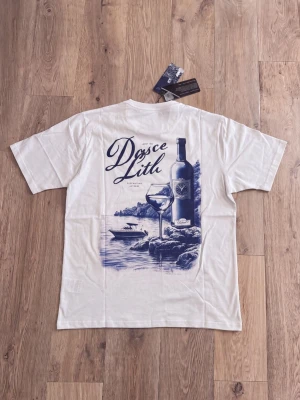 Vit t-shirt med tryck från Marcel - Vit t-shirt från Marcel med ett stort blått tryck på ryggen som visar en vinflaska, ett glas och havsmotiv samt texten 'Doce Lito'. Framtill finns en liten broderad logga på bröstet. Klassisk passform och mjukt bomullsmaterial.