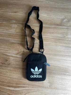Svart axelväska från Adidas - Snygg svart axelväska från Adidas med vit logga framtill. Väskan har justerbar axelrem, dragkedja och är tillverkad i slitstarkt tygmaterial. Perfekt för att bära det viktigaste och ger en sportig vibe till din outfit.