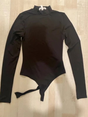 Svart långärmad body med polokrage - Snygg svart body med lång ärm och hög polokrage. Tillverkad i stretchigt, mjukt material som sitter tight mot kroppen. Praktisk knäppning i grenen med tryckknappar. Perfekt att styla till jeans eller kjol för en clean look.