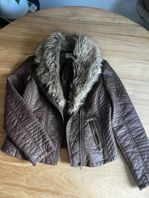 Brun skinnjacka med fuskpäls från George - Snygg brun skinnjacka från George med stor, fluffig fuskpäls runt kragen. Jackan har dragkedja framtill, två snedställda fickor med dragkedja och coola sömdetaljer på ärmar och axlar. Perfekt för dig som vill ha en edgy look.
