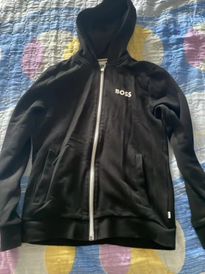Svart hoodie med dragkedja från Hugo Boss - Snygg svart hoodie från BOSS med vit dragkedja och logga på bröstet. Hoodien har huva, två fickor framtill och ribbade muddar vid ärmslut och nederkant. Perfekt för en avslappnad och stilren look.