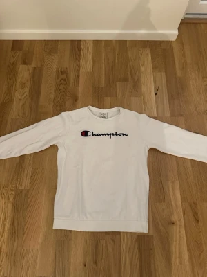 Champion Tröja - Vit långärmad tröja från Champion med klassisk logga broderad framtill i blått, rött och svart. Tröjan har rund halsringning och är tillverkad i mjukt bomullsmaterial. Perfekt för en avslappnad och sportig stil.