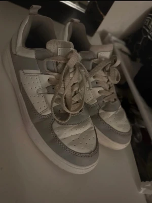 Vita och grå sneakers med perforering - Snygga sneakers i vitt och ljusgrått med perforerade detaljer på tån och sidan. Skorna har klassisk snörning och en tjock vit sula. Perfekt för dig som gillar en clean och stilren look till vardagsoutfiten.