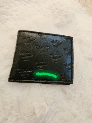 Svart plånbok med präglad logga - Snygg svart plånbok från Emporio Armani i läder med präglade loggor över hela ytan. Plånboken har flera kortfack och ett genomskinligt fack för ID eller körkort. Perfekt för dig som vill ha stilren och praktisk accessoar.