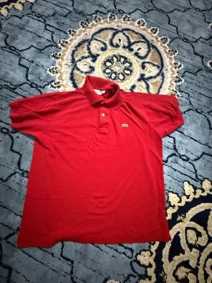 Röd pikétröja från Lacoste - Klassisk röd pikétröja från Lacoste med korta ärmar och krage. Tröjan har två knappar framtill och den ikoniska gröna krokodilloggan på bröstet. Tillverkad i bomull för en bekväm och luftig känsla.
