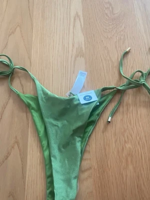 Grön bikini med knytband - Snygg grön bikini med glansig finish och smala knytband i sidorna. Bikinin har en enkel och stilren design med guldfärgade detaljer på snörena. Perfekt för strand eller poolhäng i sommar.