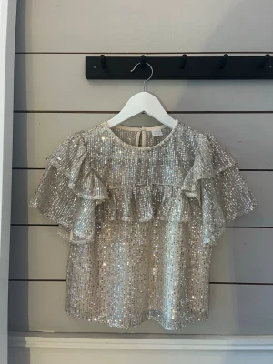 Glittrig guld  topp med volanger - Supersnygg glittrig topp i guld med korta ärmar och volangdetaljer över axlar och bröst. Toppen har rund halsringning och är täckt av paljetter som ger ett riktigt statement-utseende. Perfekt för dig som vill sticka ut och glänsa.