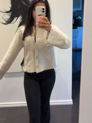 Beige stickad kofta med knappar - Mysig beige stickad kofta med långa ärmar och ribbade muddar. Koftan har en rak passform och stängs med vita knappar framtill. Perfekt för lager på lager och enkel att matcha till olika outfits.