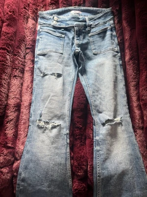 Ljusblå slitna bootcut jeans - Säljer ett par ljusblå bootcut jeans med slitningar och hål på båda benen. Jeansen har två framfickor och dubbla knappar i midjan. Perfekta för en avslappnad och trendig look.