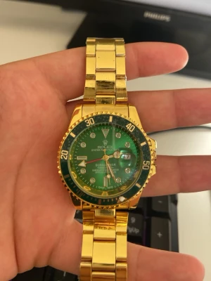  Submariner guldgrön klocka - Säljer en Submariner med grön urtavla och guldfärgat armband. Klockan har klassisk rund form, tydliga markeringar och datumfönster. Armbandet är i metall med polerad finish och boetten har en grön vridbar ring. Perfekt för dig som gillar lyxig stil.