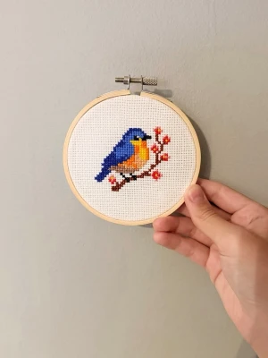Broderad tavla med blå fågel - Handgjord broderad tavla i rund träram med motiv av en blå och orange fågel som sitter på en gren med röda bär. Tavlan är sydd på vit aidaväv och ramen är i ljust trä. 