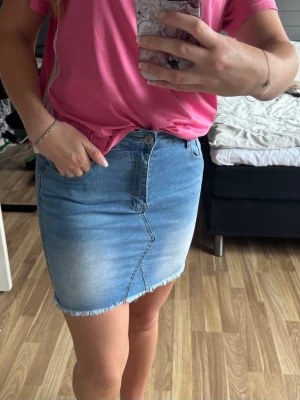 Blå jeanskjol  - Säljer en blå jeanskjol i klassisk modell med fransig kant nertill och snygga slitningar. Kjolen har normal passform, knapp och dragkedja framtill samt fickor både fram och bak. Perfekt till sommaren och enkel att matcha med olika toppar.