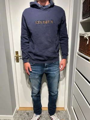 Mörkblå hoodie från Les Deux - Snygg mörkblå hoodie från Les Deux med broderad logga i guld på bröstet. Klassisk känguruficka framtill, ribbade muddar och huva med dragsko. Perfekt för en avslappnad och trendig stil.