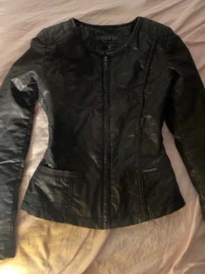 Svart skinnjacka från Guess - Snygg svart skinnjacka från Guess med figurnära passform och quiltade detaljer på axlarna. Jackan har dragkedja framtill, två fickor och diskret logga på ena fickan. Perfekt för dig som vill ha en stilren och trendig look.