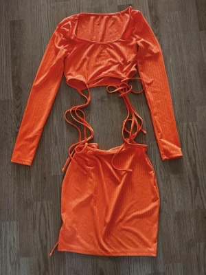Orange långärmat set med snörning - Snyggt orange set med långärmad crop top och matchande kjol. Toppen har rund halsringning och långa ärmar, båda delarna har trendiga snörningsdetaljer i midjan. Materialet är stretchigt och glansigt, perfekt för en statement-look.