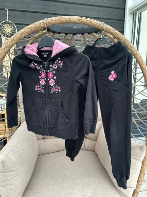 Mjukisset med broderade blommor - Mysigt svart mjukisset med dragkedja och fodrad huva med rosa fluffigt tyg. Setet har broderade rosa och vita blommor på både tröjan och byxan.