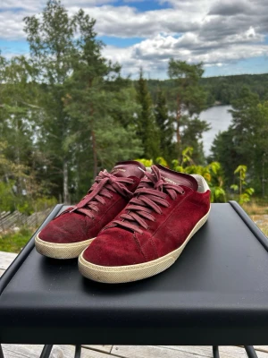 Vinröda Saint Laurent sneakers i mocka - Snygga vinröda sneakers från Saint Laurent i mjuk mocka med vita detaljer på hälen och vit sula. Klassisk låg modell med matchande vinröda skosnören och diskret logga på plösen. Perfekta för dig som gillar stilrena och exklusiva sneakers.