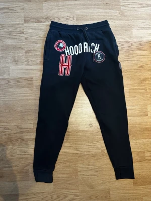 Svarta mjukisbyxor med tryck - Svarta mjukisbyxor med flera broderade och tryckta detaljer på framsidan, bland annat texten 'HOODRICH' och stora röda H. Byxorna har resår och snörning i midjan samt muddar vid benslut. Perfekta för en avslappnad och cool look.