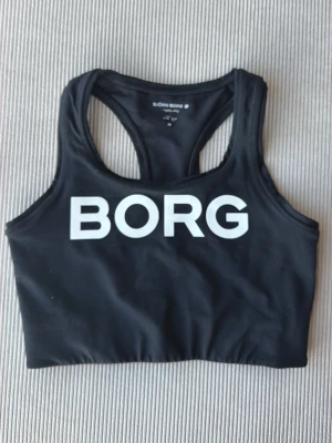 Svart sport-bh från Björn Borg - Snygg svart sport-bh från Björn Borg med bred resår och stor vit logga framtill. Modellen har brottarrygg och är tillverkad i ett stretchigt funktionsmaterial som passar perfekt till träning och gym.