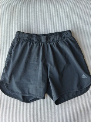 Svarta träningsshorts från Adidas - Snygga svarta träningsshorts från Adidas med elastisk midja och diskret logga på benet. Shortsen har en sportig passform och är tillverkade i lätt syntetmaterial som andas. Perfekta för löpning eller gymmet.