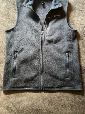 Fleece väst patagonia - Snygg mörkblå fleeceväst från patagonia med hel dragkedja framtill och tre fickor med dragkedja. Västen har en hög krage och en enkel, stilren design som passar till många olika outfits.