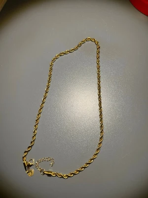 Guld cordellhalsband 60cm  - Snyggt halsband i guldtonad metall med tvinnad kedja och ett litet hjärthänge vid låset. Halsbandet har en justerbar kedja för perfekt passform och ger en elegant touch till din stil. Perfekt för dig som gillar klassiska accessoarer med en twist