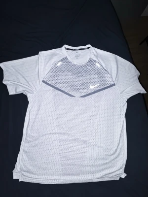 Vit sporttröja från Nike - Snygg vit sporttröja från Nike med grått mönster på bröstet och ryggen. Tröjan har korta ärmar, rund halsringning och är tillverkad i ett lätt och ventilerande material som passar perfekt för träning. Diskret Nike-logga på bröstet. /////    Passar XL    //////