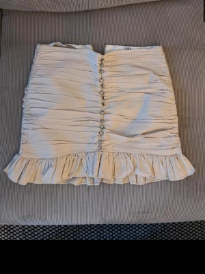 Sötaste kjolen!! - Supersöt beige kortkjol med volangkant och dekorativa knappar framtill. Kjolen har rynkade detaljer som ger en snygg struktur och feminin look. Perfekt att styla med en enkel topp eller stickad tröja. Från Zara, märkt med L men skulle säga att passar s o m bättre💕