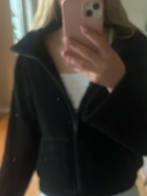Svart teddyjacka med dragkedja - Mysig svart teddyjacka med hög krage och dragkedja framtill. Jackan har en fluffig, mjuk yta och två fickor på sidorna. Perfekt för kyliga dagar och enkel att matcha med olika outfits.