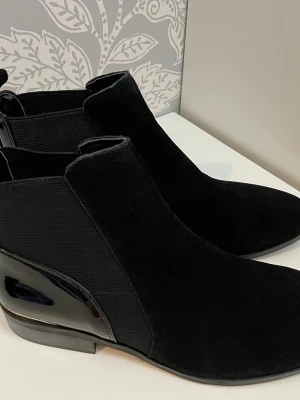 Eleganta snygga Chelsea boots från River Island, stl 40 oanvända  - Snygga svarta Chelsea boots med klassisk resår på sidorna och cool lackdetalj vid hälen. Bootsens ovandel är i mockaliknande material och de har en låg klack. Perfekta för dig som vill ha en stilren och trendig look. Stl 40, äkta mocka 