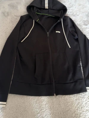 Svart hoodie med dragkedja från BOSS - Snygg svart hoodie från BOSS med dragkedja framtill och vita detaljer på huva och ärmslut. Hoodien har fickor på magen och en diskret BOSS-logga på bröstet. Insidan av huvan har en grön kant som ger en fräsch kontrast. Perfekt för en chill och stilren look. Från Johnells orginal pris för hela dressen var Ca 3400 