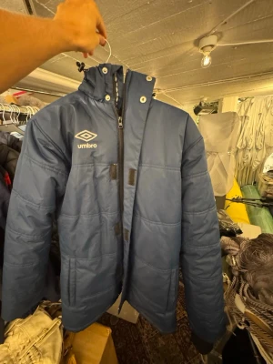 Blå dunjacka från Umbro med huva - Snygg blå dunjacka från Umbro med avtagbar huva och dragkedja framtill. Jackan har Umbro-logga på bröstet och ärmen, samt praktiska fickor. Perfekt för kalla vinterdagar och riktigt varm tack vare det tjocka materialet. Storlek passar M också. Finns att hämta i Gröndal.