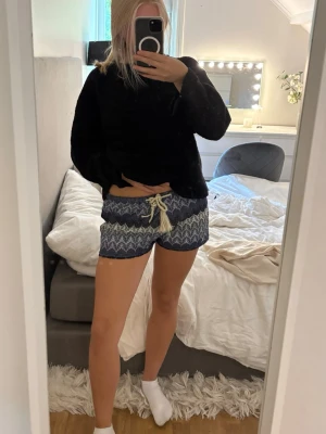 Blå mönstrade shorts med snörning - Säljer ett par blå shorts med vitt och grått zigzagmönster och dekorativ snörning i midjan. Shortsen har en avslappnad passform och är perfekta för varma dagar. Materialet känns mjukt och bekvämt mot huden.