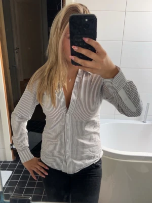 Vit och grårandig skjorta - Säljer en vit skjorta med tunna grå ränder. Skjortan har lång ärm, knappar framtill och klassisk krage. Passformen är normal och materialet känns som bomull, vilket gör den bekväm och enkel att styla till olika outfits.